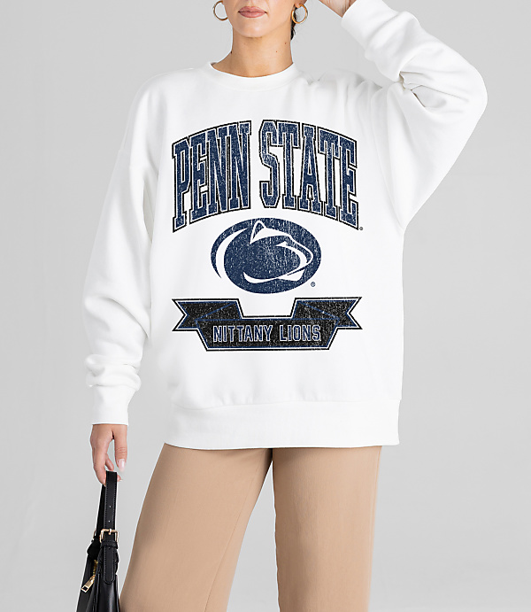A woman in a white Penn State sweatshirt and tan pants stands against a plain gray background, holding a black purse.