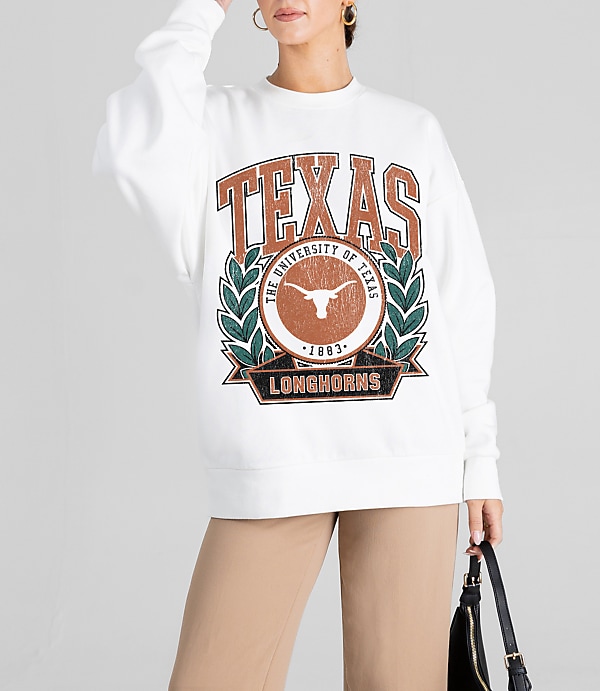 A woman in a white sweatshirt with a University of Texas Longhorns graphic stands against a plain gray background, holding a black purse.