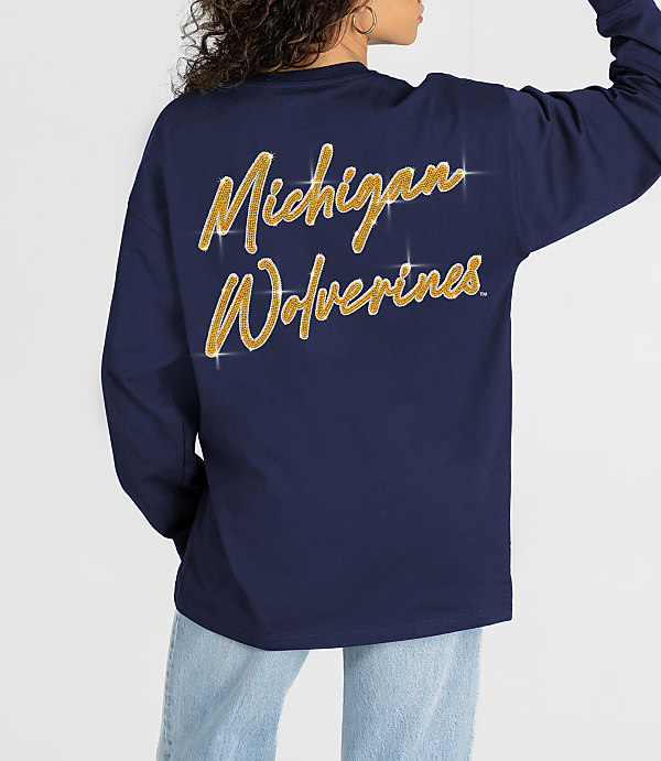 A person in a navy long-sleeved shirt with "Michigan Wolverines" in gold glitter on the back stands against a plain white background.