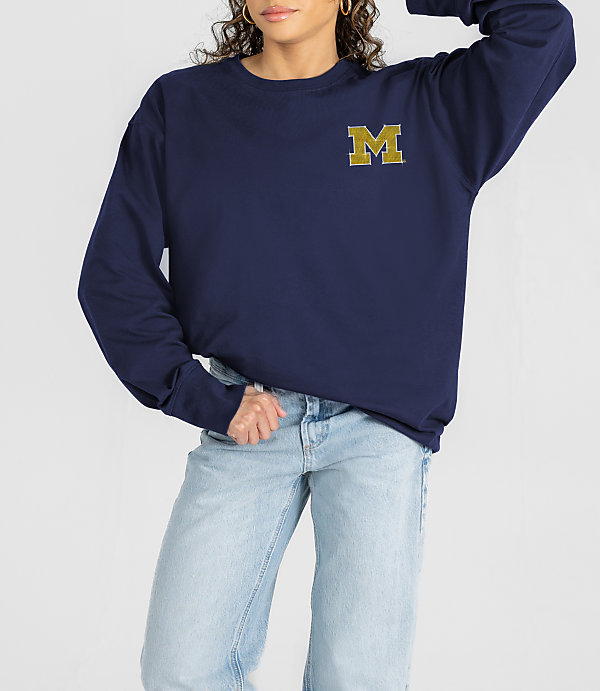 A woman in a navy sweatshirt with a gold "M" logo and light-wash jeans stands against a plain white background.