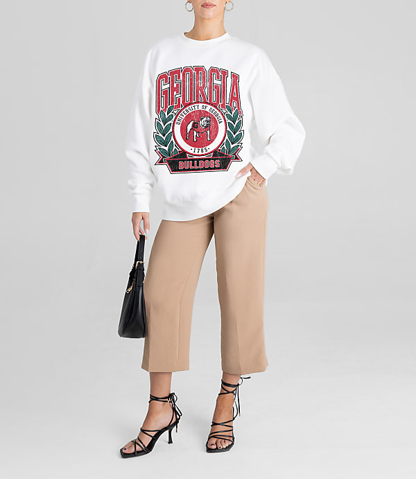 A woman in a white sweatshirt and tan pants stands against a plain white background, holding a black purse.