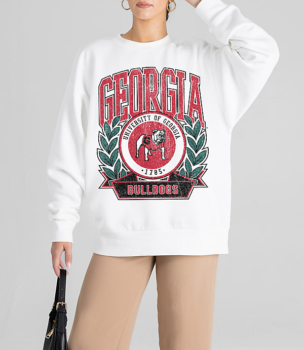 A person in a white sweatshirt with a distressed "Georgia Bulldogs" logo stands against a plain background, holding a black purse.