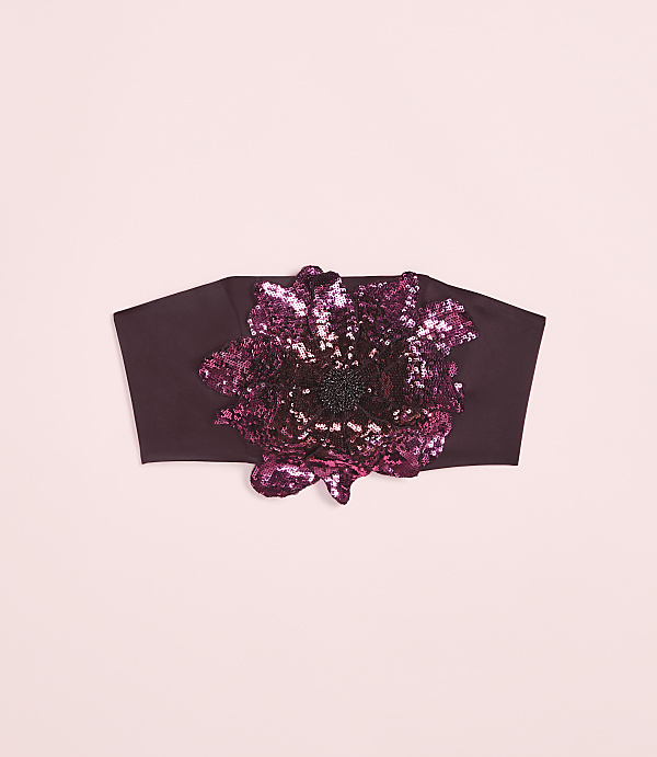 A dark plum headband with a large, sequined floral embellishment sits against a soft pink background.