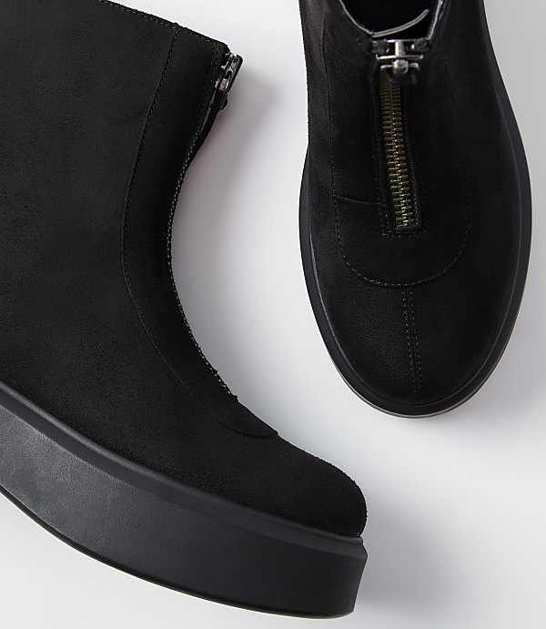 A pair of black suede ankle boots with front zippers rests on a white surface.