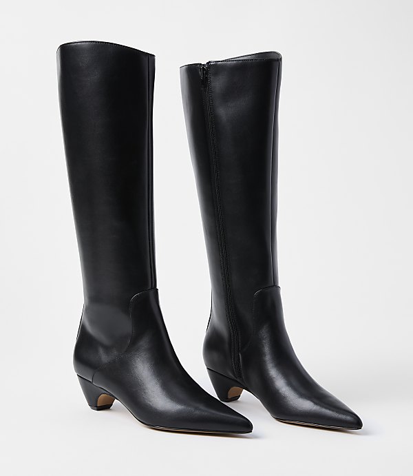 A pair of black leather knee-high boots with pointed toes and low heels stand on a white background.