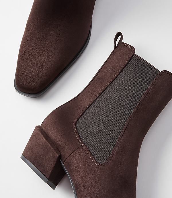 A pair of brown suede Chelsea boots with block heels are arranged on a white background, with one boot in the foreground and the other partially visible in the background.