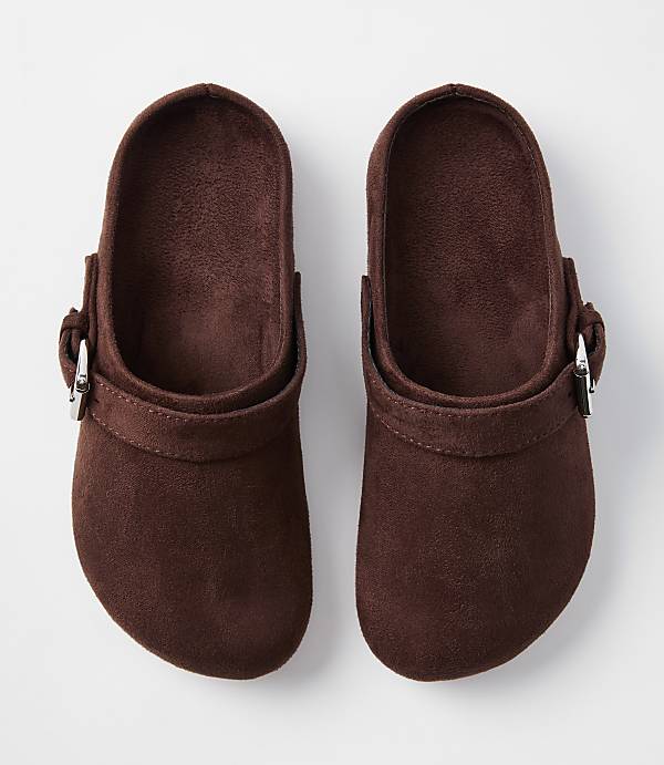 A pair of brown suede clogs with silver buckles rests on a plain white background.
