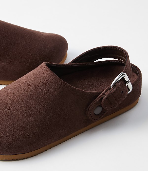 A pair of brown suede clogs with a silver buckle rests on a white surface, with another clog partially visible in the background.