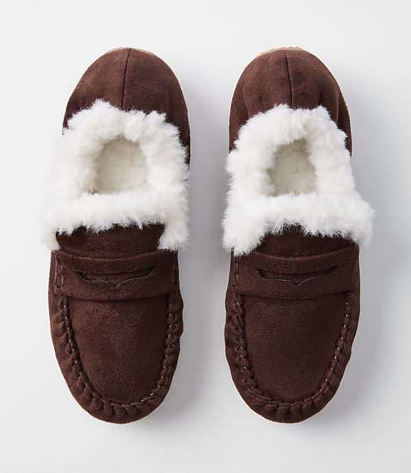 A pair of brown suede moccasin slippers with white fluffy lining are displayed against a plain white background.