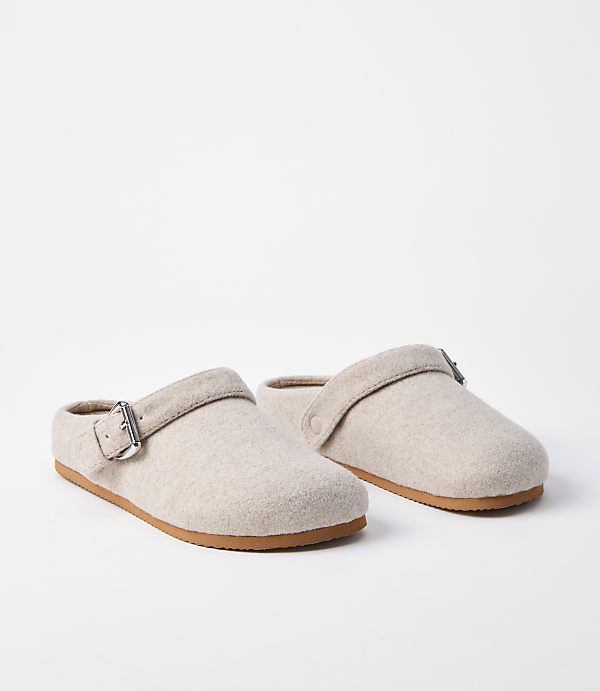 A pair of light beige felt clogs with buckle straps rests on a white surface, with a plain white background behind them.