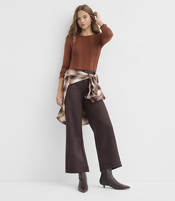 A woman in a brown sweater, dark wide-leg pants, and ankle boots stands against a plain white background, with a plaid shirt tied around her waist.