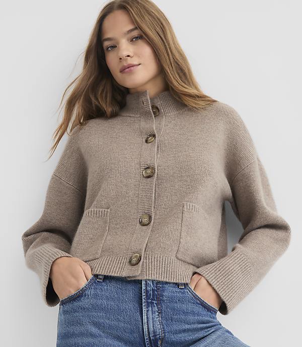 A woman in a beige cardigan and blue jeans stands against a plain white background.