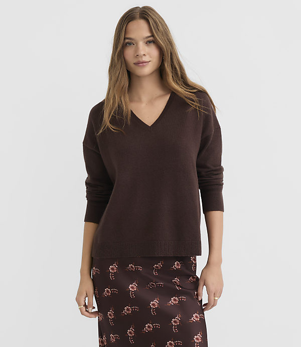 A woman in a brown v-neck sweater and floral skirt stands against a plain gray background.