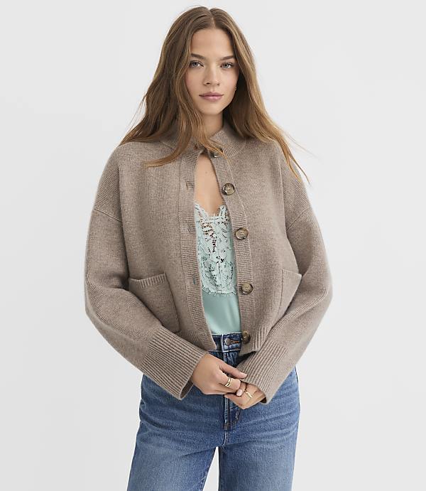A young woman in a beige cardigan and blue jeans stands against a plain white background.