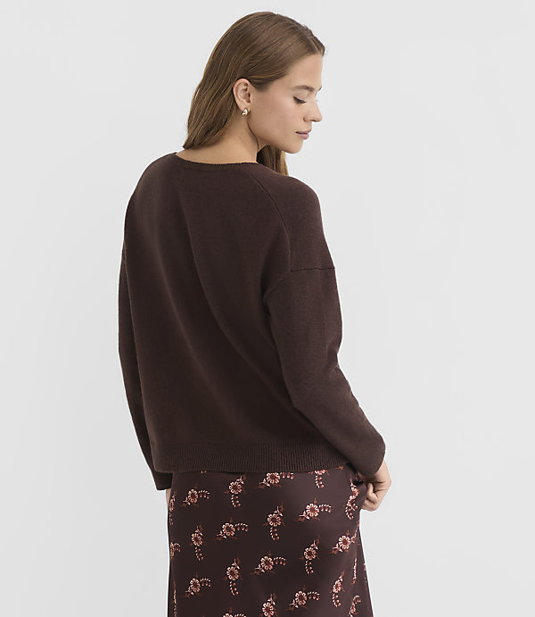 A woman in a brown sweater and floral skirt stands against a plain gray background.