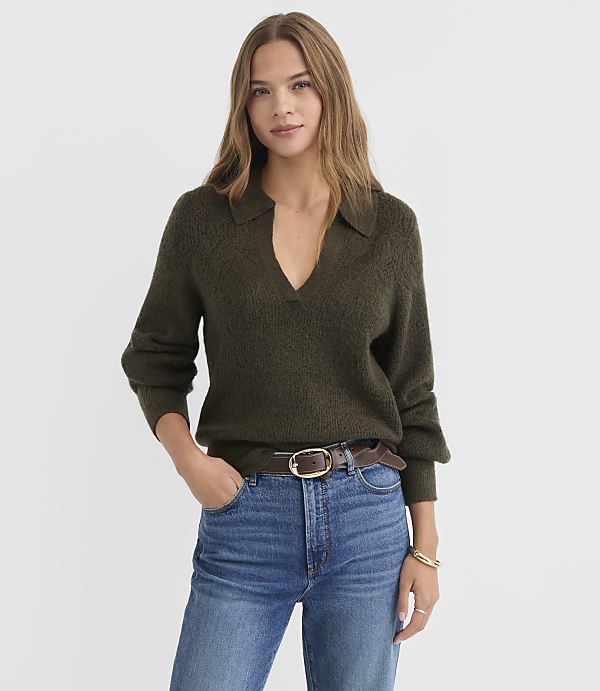 A woman in a dark green collared sweater and blue jeans stands against a plain white background.