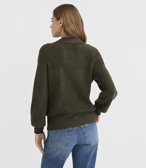 A woman in a textured olive green sweater and blue jeans stands against a plain, light gray background.