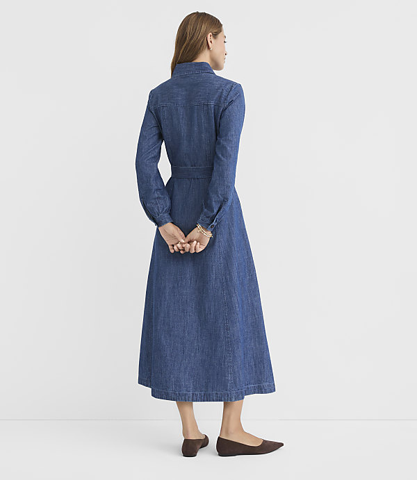 A woman in a denim dress stands with her hands clasped behind her back against a plain white background.