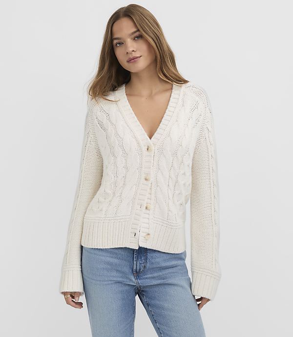 A woman in a cream cable-knit cardigan and blue jeans stands against a plain white background.