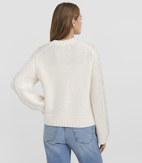A woman in a cream cable-knit sweater and blue jeans stands against a plain white background.
