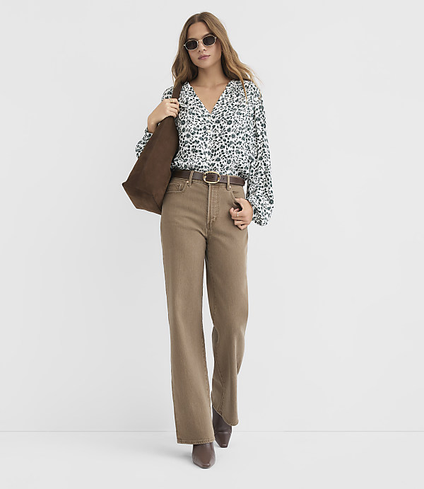 A woman in a floral blouse and wide-leg brown pants stands against a plain white background, carrying a brown tote bag.