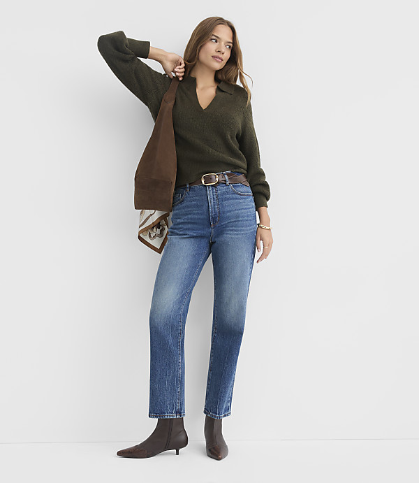 A woman in a green sweater and blue jeans stands against a plain white background, holding a brown bag over her shoulder.