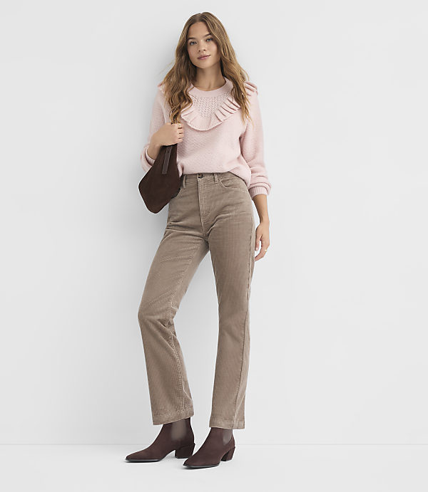 A woman in a pink ruffled sweater and brown corduroy pants stands against a plain white background, holding a dark brown purse.