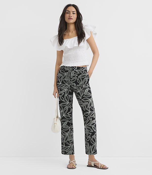 A woman in a white ruffled top and black leaf-patterned pants stands against a plain white background, holding a small white purse.