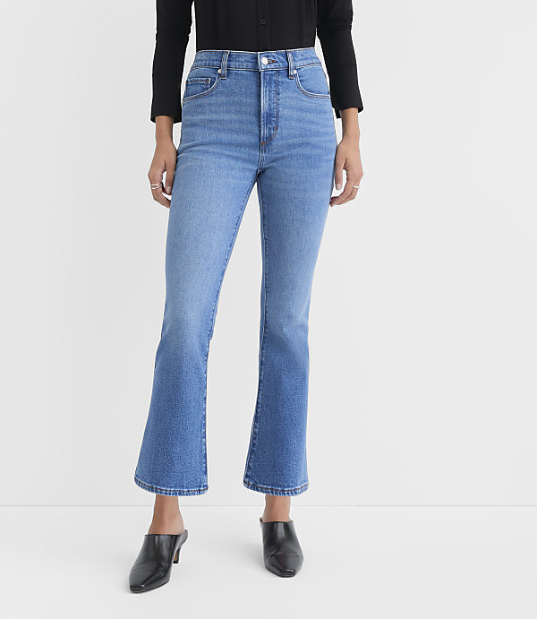 A person wearing flared blue jeans and black heeled mules stands against a plain white background.