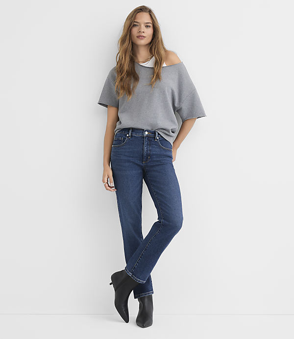 A woman in a gray off-the-shoulder top and dark wash jeans stands against a plain white background.