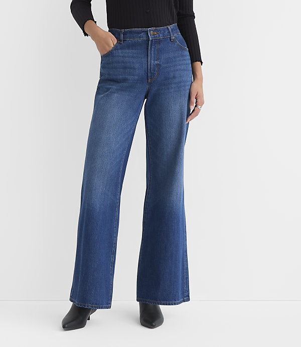 A person wearing wide-leg blue jeans and black heeled boots stands against a plain white background.