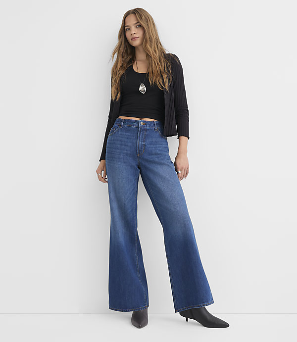 A young woman in a black top, cardigan, and wide-leg jeans stands against a plain white background.