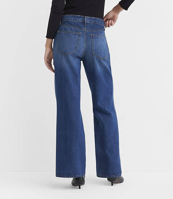 A person wearing wide-leg blue jeans and black heels stands against a plain white background.