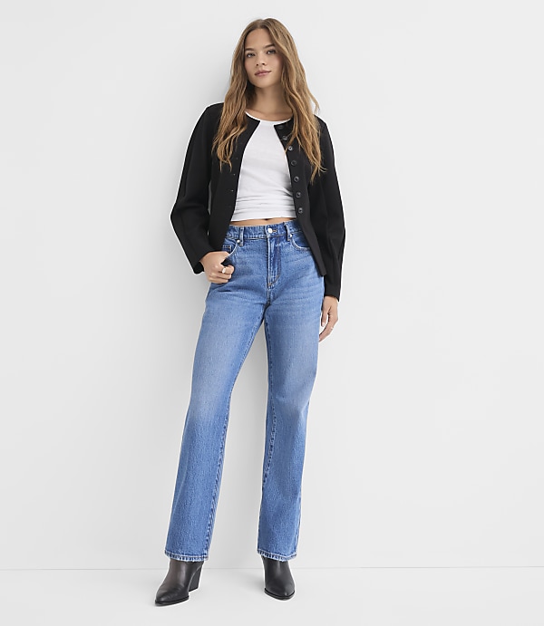 A woman in a white crop top, black jacket, and blue jeans stands against a plain white background.