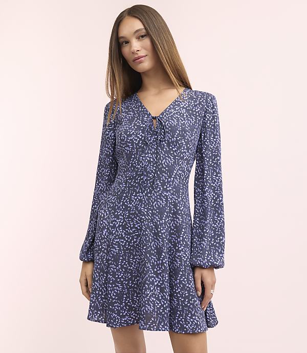 A woman in a blue floral dress stands against a soft pink background.