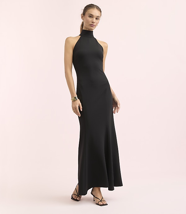 A woman in a black halter neck gown stands against a soft pink background.