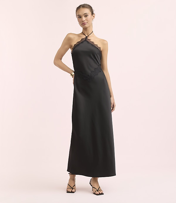 A woman in a black halter dress with lace trim stands against a soft pink background.
