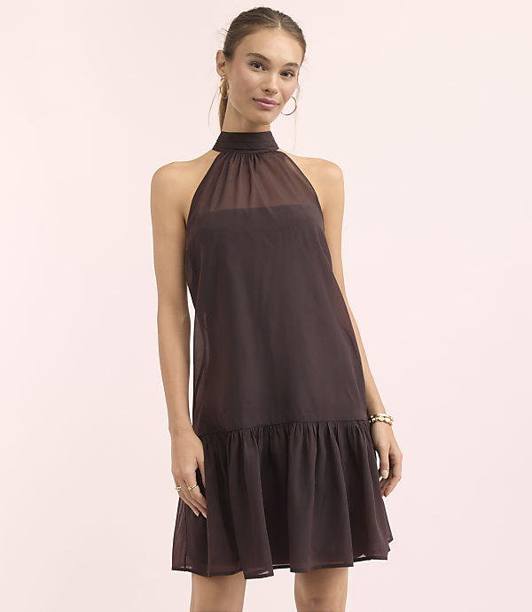 A woman in a dark brown halter dress stands against a soft pink background.
