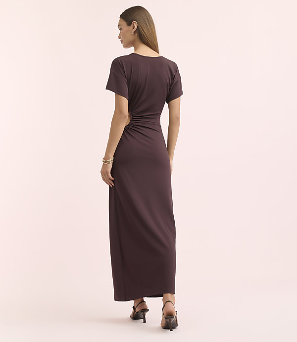 A woman in a dark purple, ruched maxi dress stands against a soft pink background.