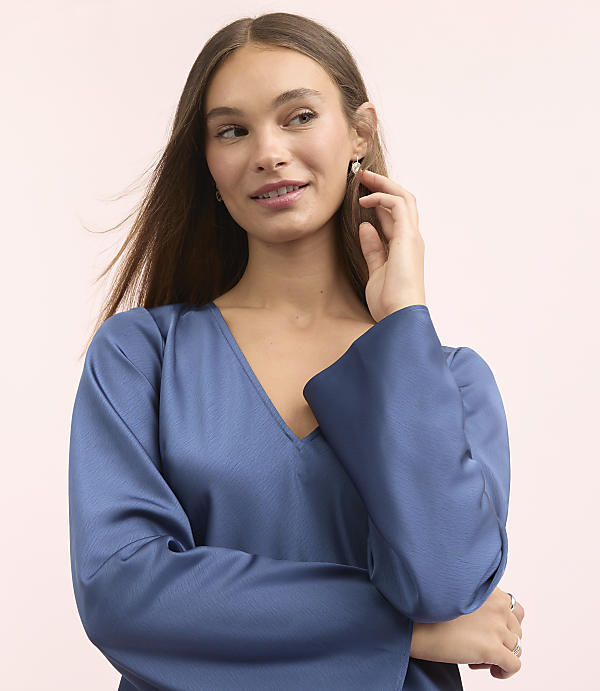A young woman with long brown hair and a blue satin top poses with her hand near her ear against a soft pink background.