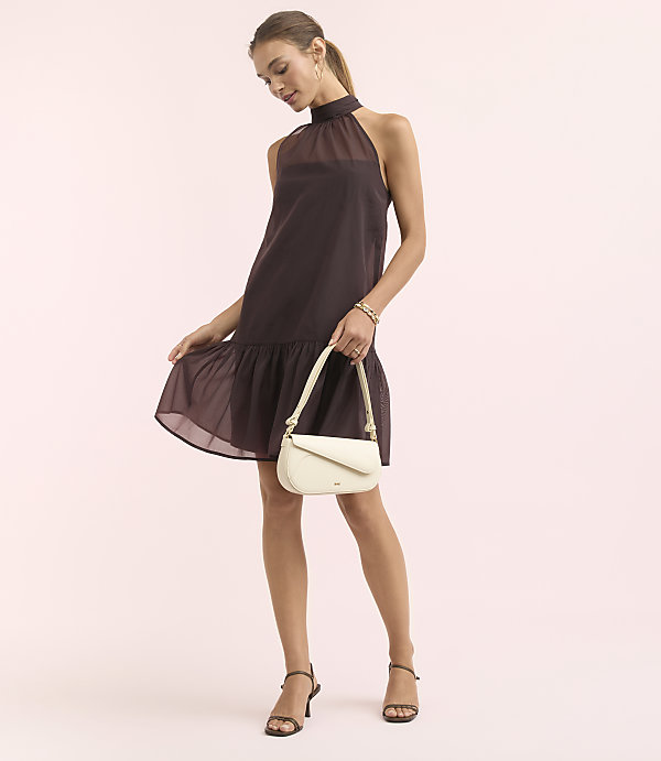 A woman in a dark purple halter dress and cream handbag stands against a soft pink background.