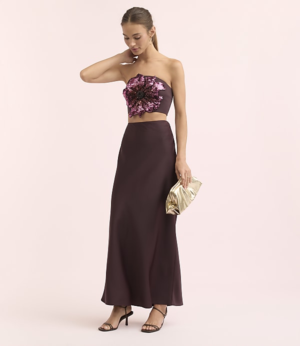 A woman in a strapless floral top and a long skirt poses with a gold clutch against a soft pink background.