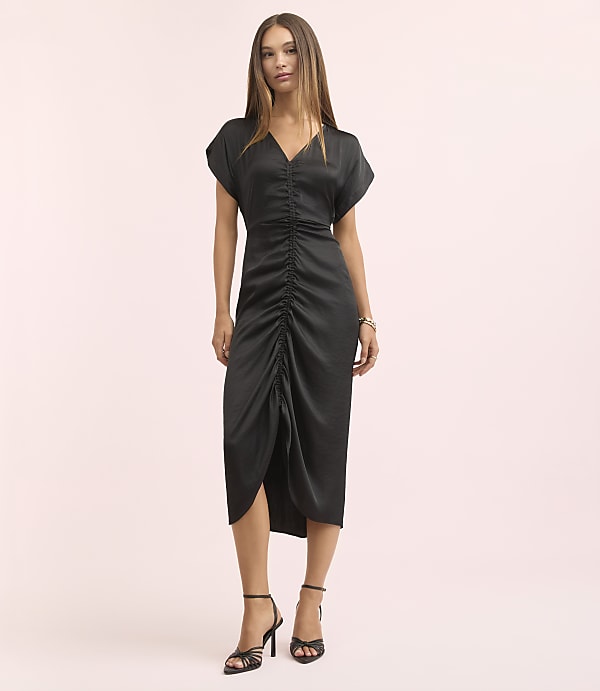 A woman in a black ruched dress and strappy heels stands against a soft pink background.