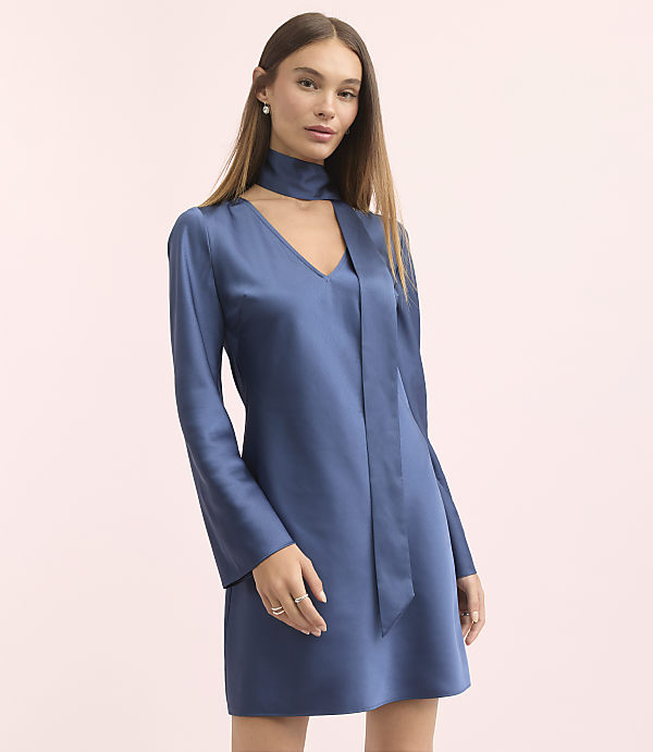 A woman in a blue satin dress with bell sleeves and a scarf neckline stands against a soft pink background.