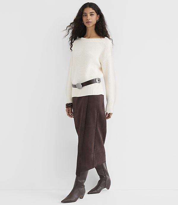 A woman in a cream sweater and brown skirt stands against a plain white background.