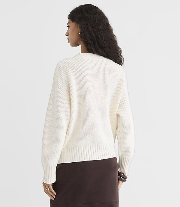 A woman in a cream-colored sweater and brown skirt stands against a plain white background.