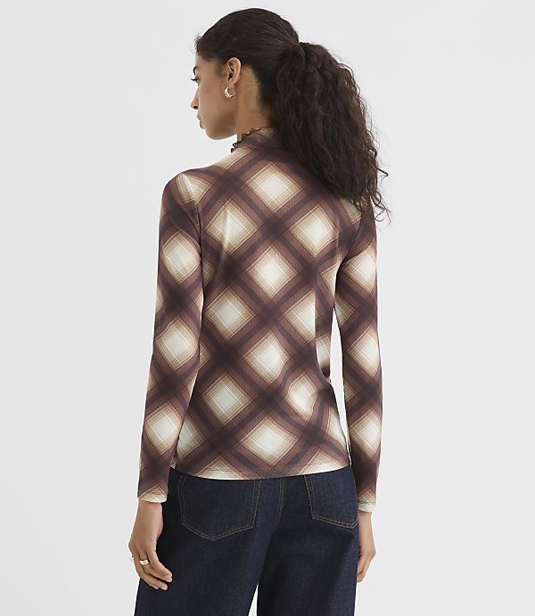 A woman in a brown plaid turtleneck and dark jeans stands against a plain white background.