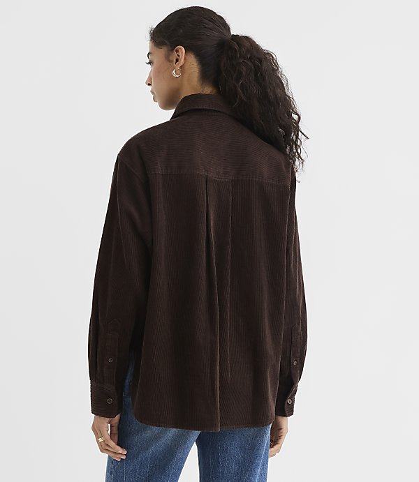 A woman in a brown corduroy shirt and blue jeans stands against a plain white background.