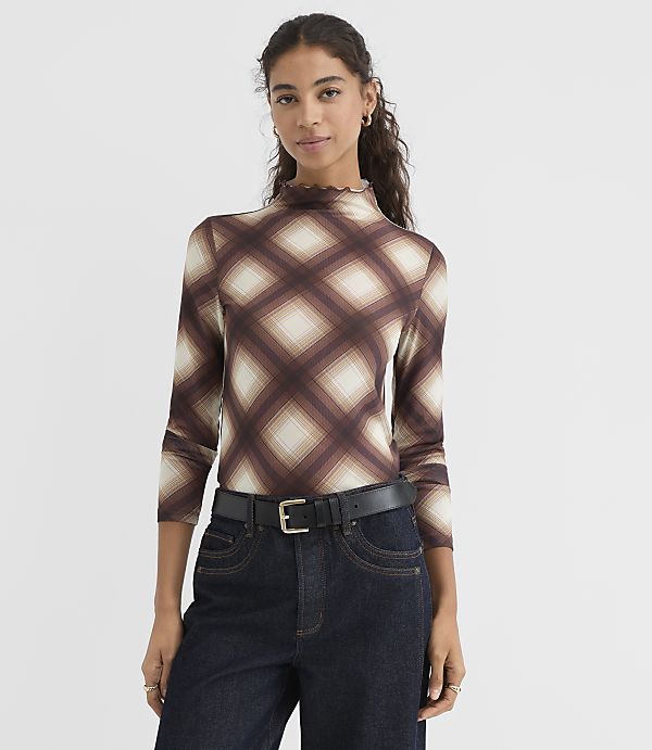 A woman in a brown plaid turtleneck and dark jeans stands against a plain white background.