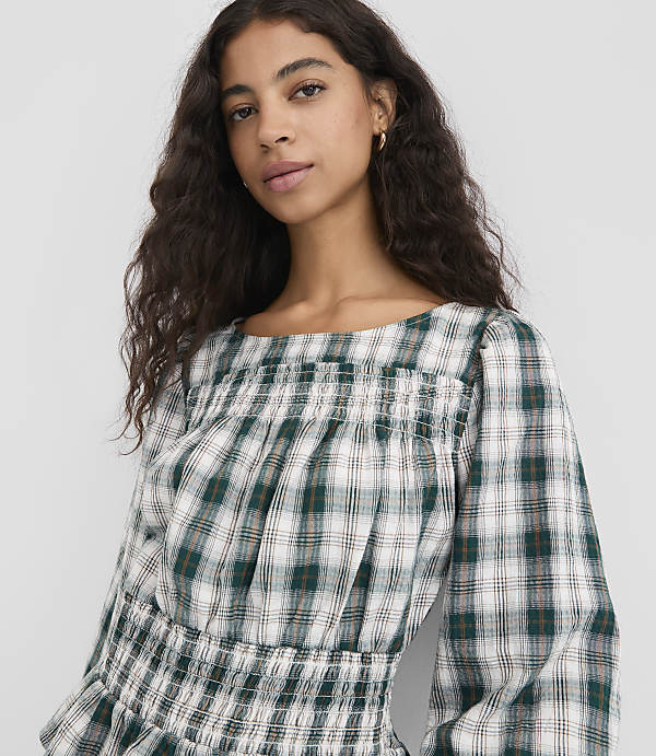 A young woman with dark curly hair wears a green and white plaid smocked top, with a plain white background.